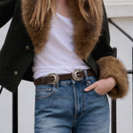 A person wearing a dark green jacket with a brown fur collar and cuffs, a white tank top, and blue jeans with a brown belt featuring large silver concho-style buckles.