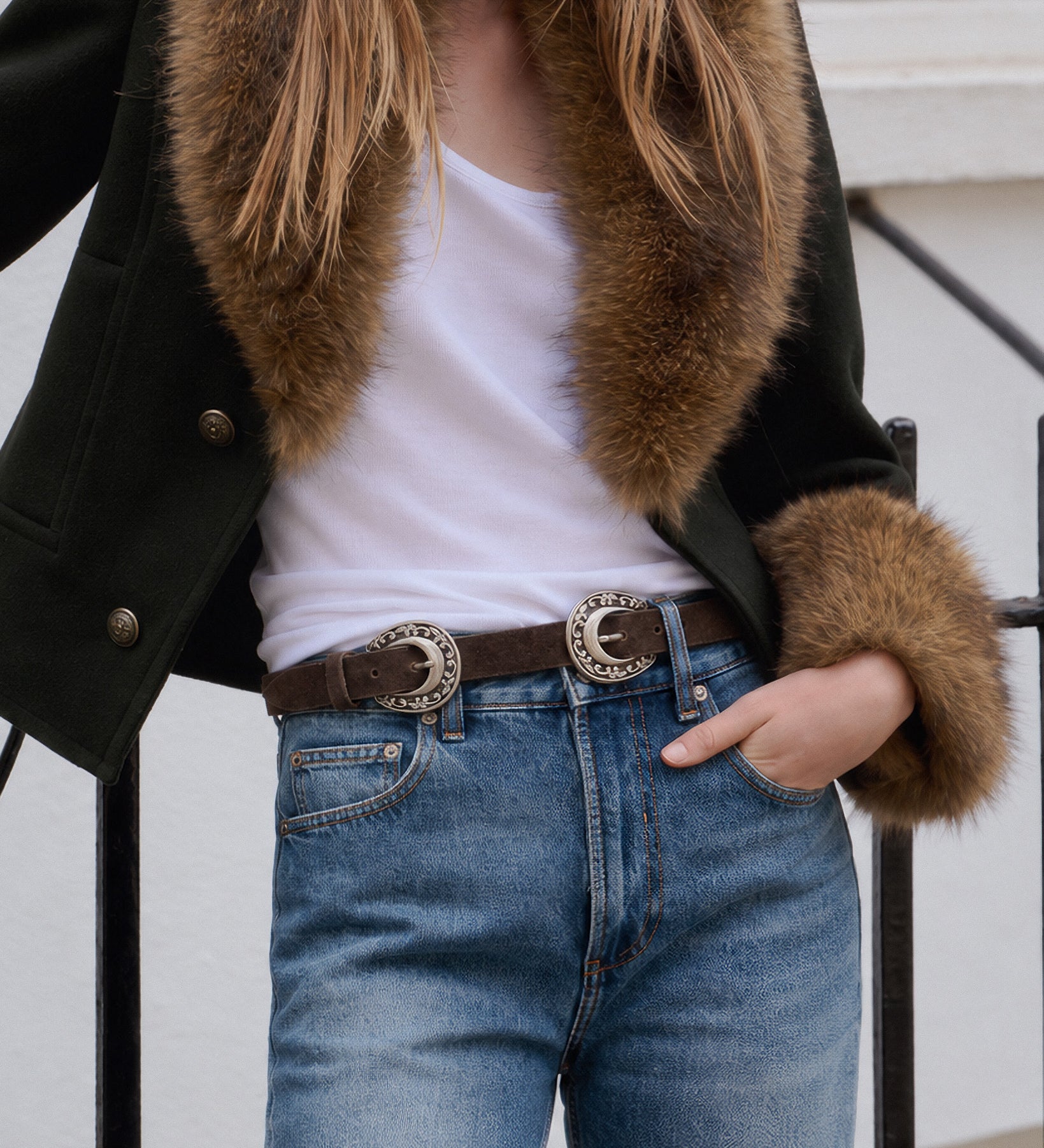 A person wearing a dark green jacket with a brown fur collar and cuffs, a white tank top, and blue jeans with a brown belt featuring large silver concho-style buckles.
