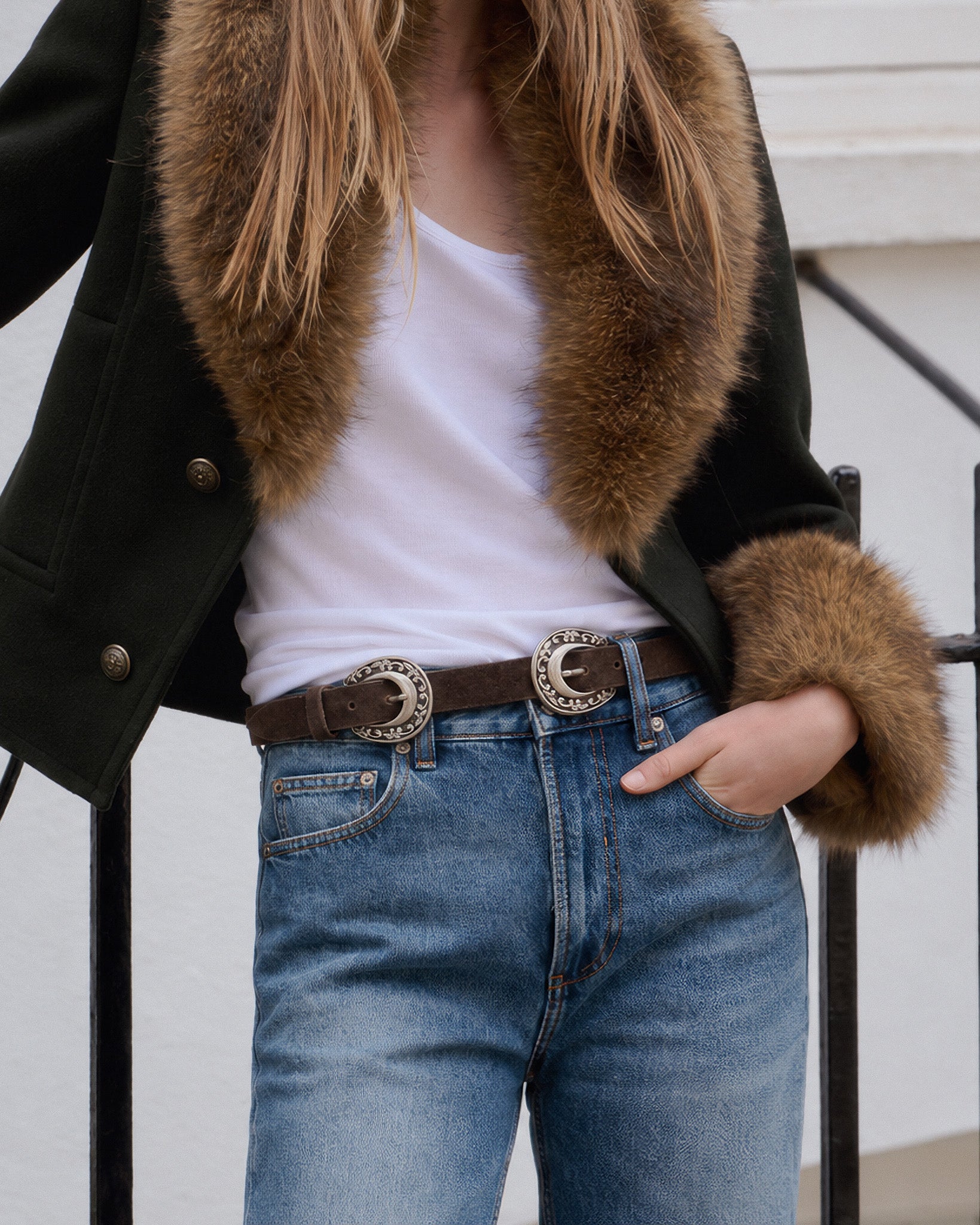 A person wearing a dark green jacket with a brown fur collar and cuffs, a white tank top, and blue jeans with a brown belt featuring large silver concho-style buckles.