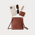 A brown leather crossbody bag with gold-tone hardware is displayed in a flat-lay arrangement on a light gray background, surrounded by lifestyle accessories including a smartphone, sunglasses, lipstick, and a cardholder.