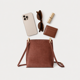 A brown leather crossbody bag with gold-tone hardware is displayed in a flat-lay arrangement on a light gray background, surrounded by lifestyle accessories including a smartphone, sunglasses, lipstick, and a cardholder.