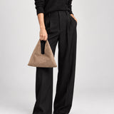 A model wearing black wide-leg trousers, a black sweater, and black pointed shoes, holding a brown suede triangular handbag with a black handle against a light gray studio background.
