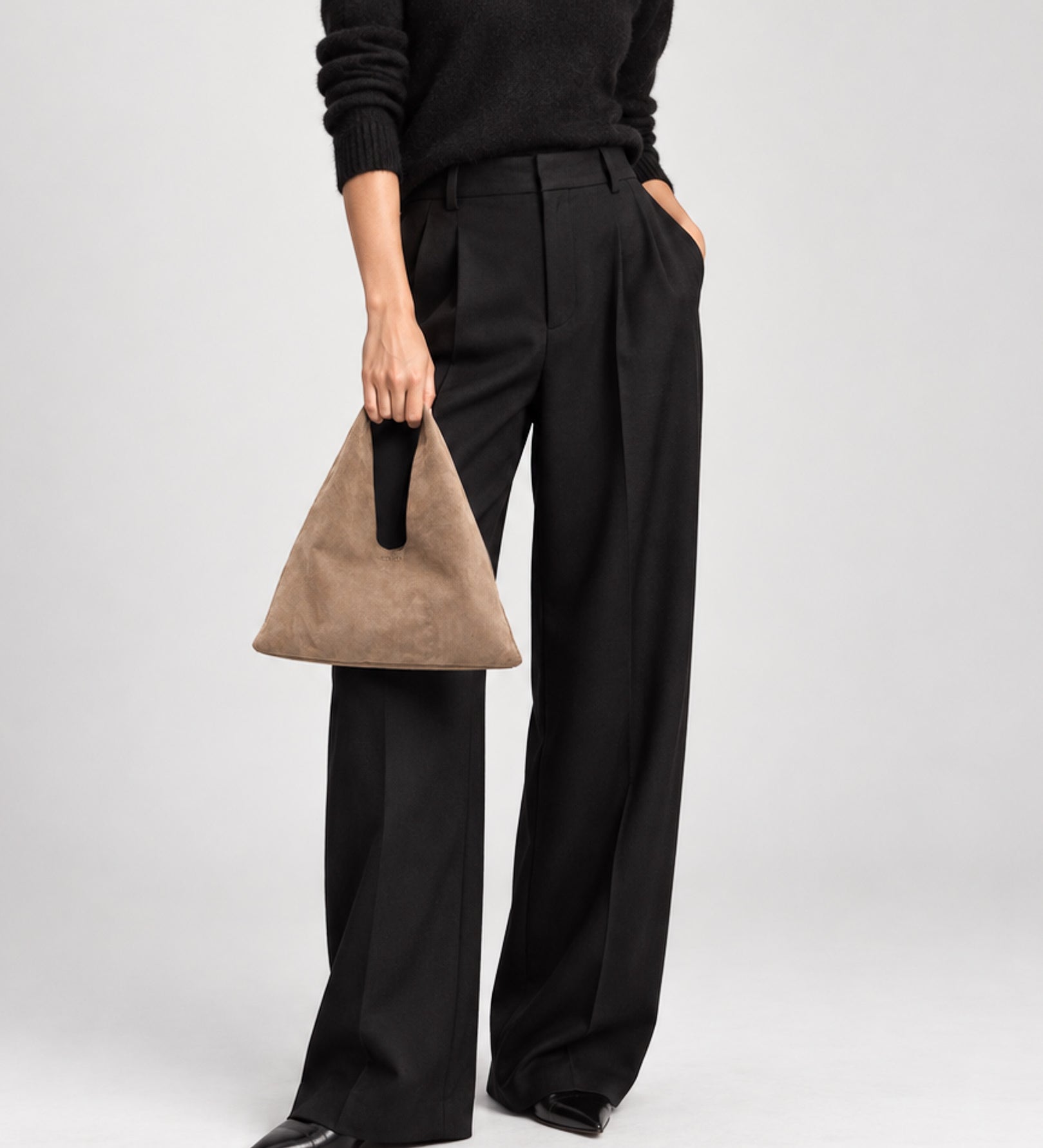 A model wearing black wide-leg trousers, a black sweater, and black pointed shoes, holding a brown suede triangular handbag with a black handle against a light gray studio background.