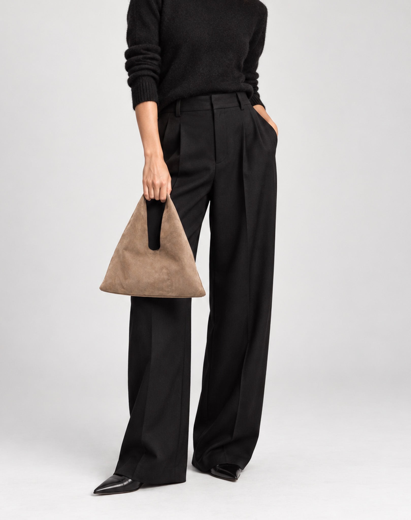 A model wearing black wide-leg trousers, a black sweater, and black pointed shoes, holding a brown suede triangular handbag with a black handle against a light gray studio background.