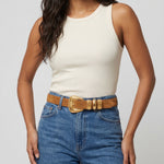 Woman wearing a white tank top and blue jeans with a brown belt on a light gray background